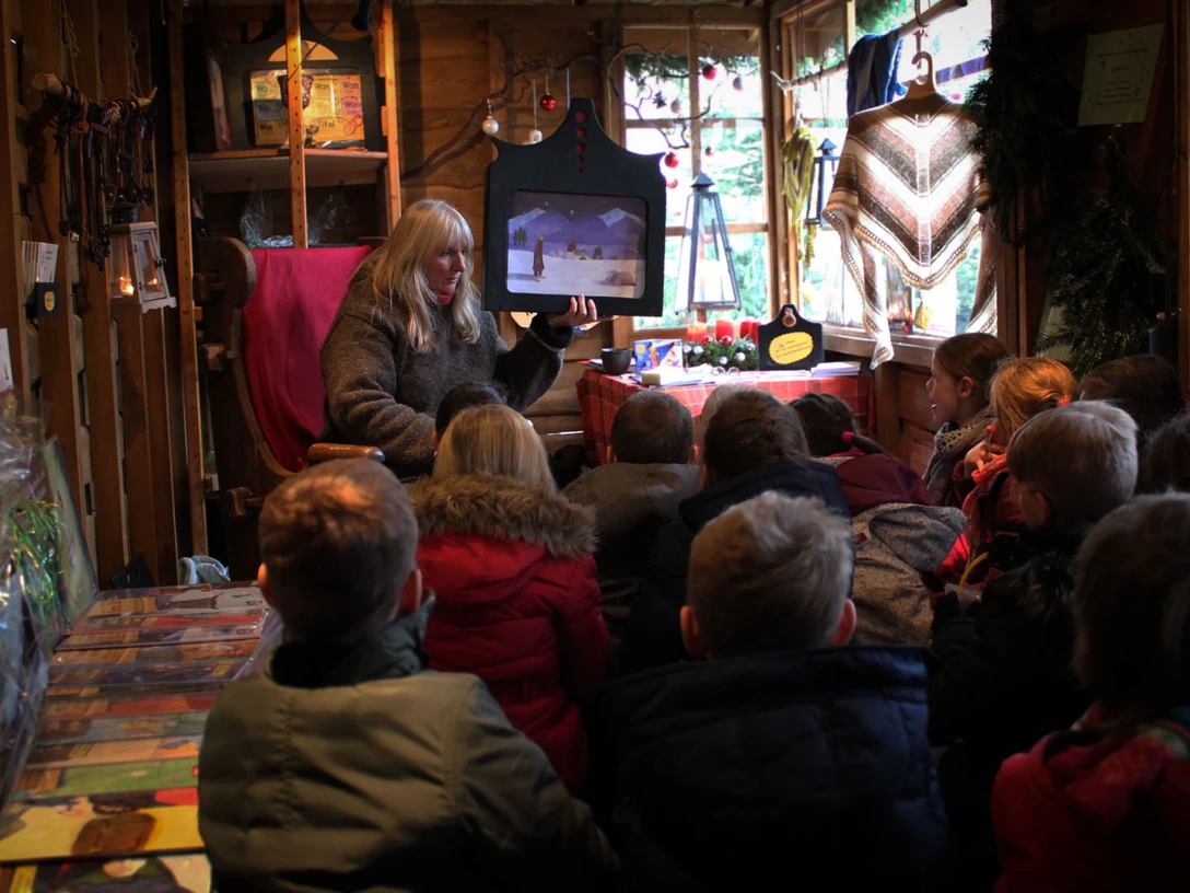 Erzählwerkstatt auf dem Weihnachtsmarkt Eine Frau liest einer Gruppe von Kindern in einer hölzernen Hütte auf dem Weihnachtsmarkt vor.A woman reads to a group of children in a wooden hut at the Christmas market.En kvinde læser op for en gruppe børn i en træhytte på julemarkedet.Een vrouw leest voor aan een groep kinderen in een houten hut op de kerstmarkt.