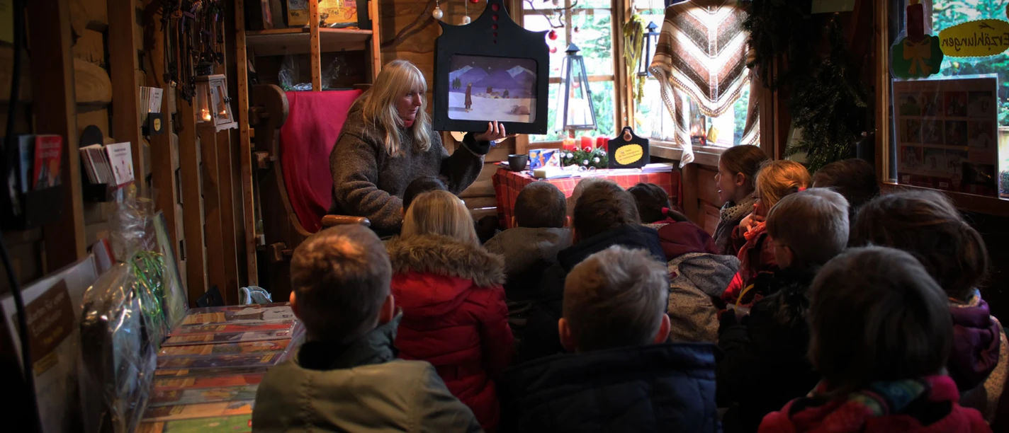 Een vrouw leest voor aan een groep kinderen in een houten hut op de kerstmarkt.