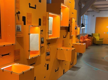 Geschichte-des-Ankommens (2).jpg Orangefarbene interaktive Ausstellungswände mit beleuchteten Schaukästen in modernem Design.