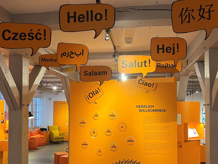Geschichte-des-Ankommens.jpg Einladende Ausstellung mit Willkommensgrüßen in verschiedenen Sprachen.