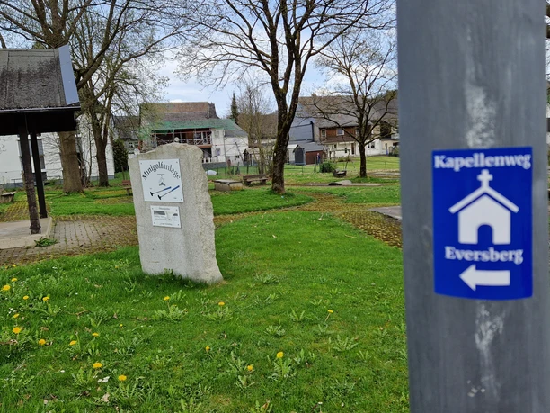 Kapellenweg Eversberg am Minigolfplatz Ein grüner Platz mit Wegweiser und Ruhebank, im Hintergrund ein kleiner Pavillon.