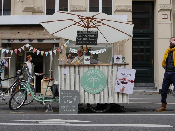 Ein mobiler Verkaufsstand auf einem Bürgersteig mit einer Person hinter einem Tresen, Fahrräder und Passanten.