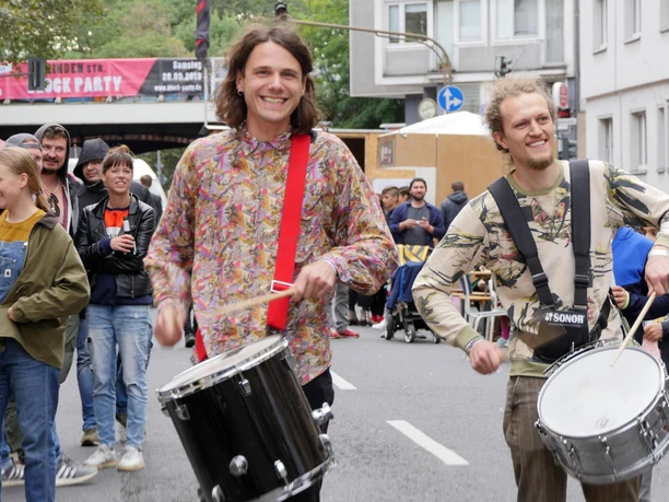Zwei Männer in bunten Hemden spielen lächelnd Trommeln bei einem Straßenfest.