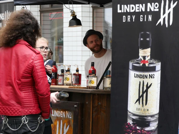 Block Party A salesman in a hat stands behind a stand selling Linden No. 5 gin, attended by two people.