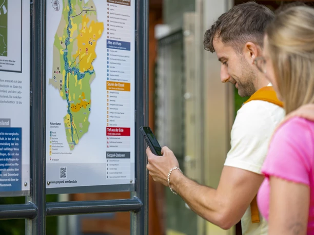 Geopark-Tafel beim Koloss von Hüven – Radfahren Hümmling ©Emsland Tourismus GmbH (4).jpg Zwei Personen betrachten eine Infotafel des Geoparks Emsland und scannen mit dem Smartphone einen QR-Code.