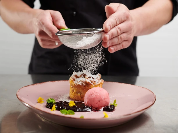 A chef sprinkles a delicious dessert with powdered sugar, accompanied by sorbet and berries on a plate.