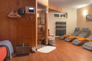 Saunaruimte met houten lambrisering: ligstoelen met oranje bekleding, houten deur naar de sauna aan de linkerkant, heldere verlichting.