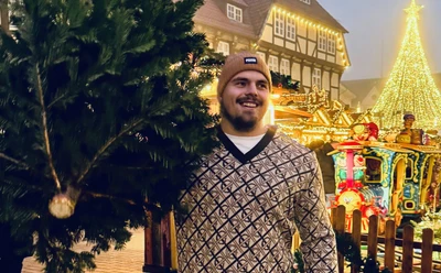 Max Dehning mit festlichem Hintergrund auf Weihnachtsmarkt, lächelnd mit Tanne.