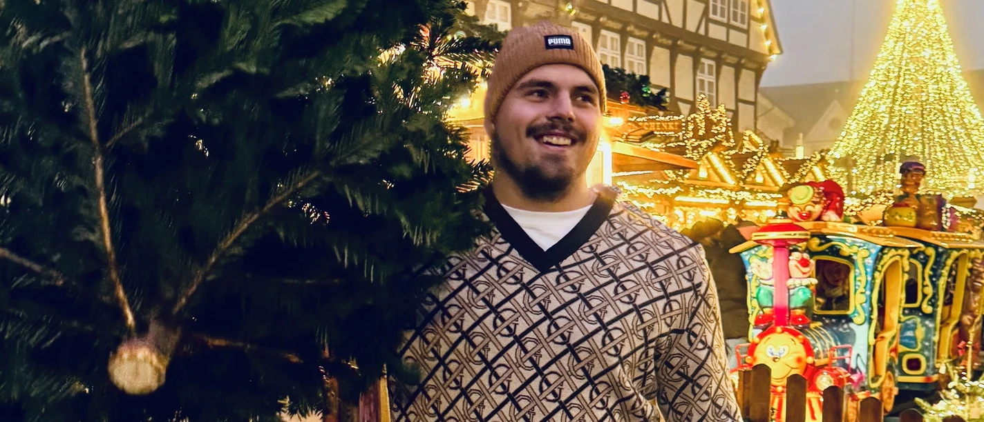 Der Gewinner von 2024: Max Dehning Max Dehning mit festlichem Hintergrund auf Weihnachtsmarkt, lächelnd mit Tanne.