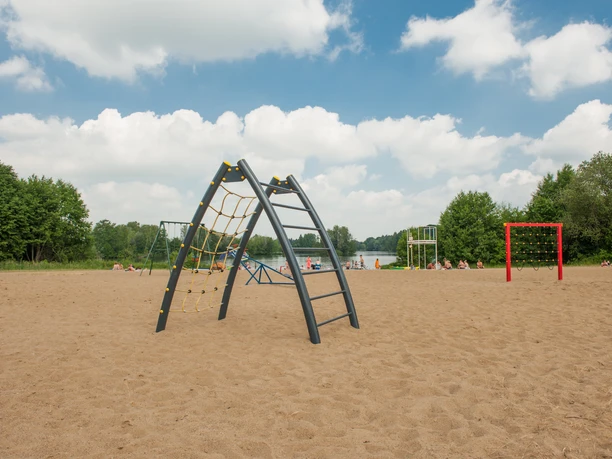 Spielplatz am Pruesssee Spielplatz am sandigen Strand mit Klettergerüst und Schaukeln, umgeben von Bäumen unter blauem Himmel.
