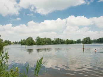 Badestelle am Prüßsee Zwei Personen schwimmen im ruhigen See, umgeben von grünen Ufern und bewölktem Himmel.