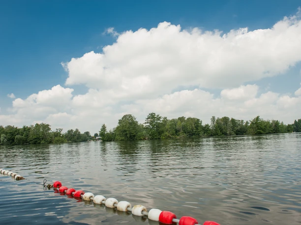 Badestelle am Prüßsee Sanftes Wasser mit grüner Insel, bewölkter Himmel darüber. Uferabsperrung mit rot-weißen Bojen.