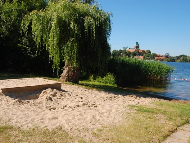 Badestelle Bäk mit Sandaksten Ein Sandstrand mit Buddelkasten führt zum Ratzeburger See mit einer Baumreihe und einem Kirchturm im Hintergrund.