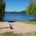 Badestelle Bäk Ein sandiger Uferbereich mit zwei herabhängenden Weidenbäumen am Seeufer und klarem blauen Himmel.