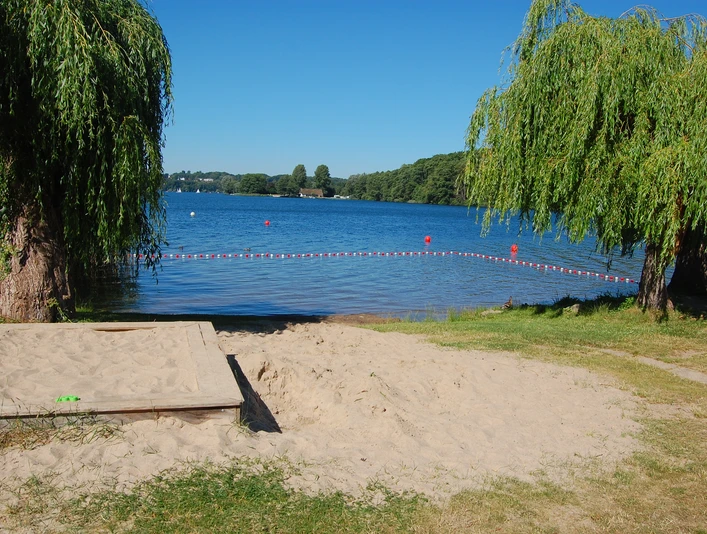 Badestelle Bäk Ein sandiger Uferbereich mit zwei herabhängenden Weidenbäumen am Seeufer und klarem blauen Himmel.
