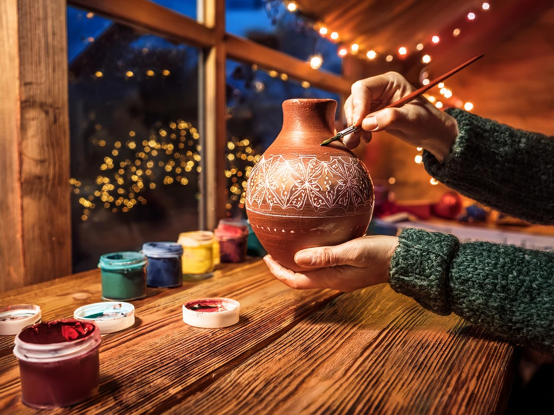 Töfperei.jpg Zwei Hände bemalen eine Vase kreativ, umgeben von Farbgläsern in einer Holzwerkstatt.Two hands creatively paint a vase surrounded by jars of paint in a wood workshop.To hænder maler kreativt en vase omgivet af farvede glas i et træværksted.Twee handen beschilderen creatief een vaas omringd door gekleurde glazen in een houtwerkplaats.