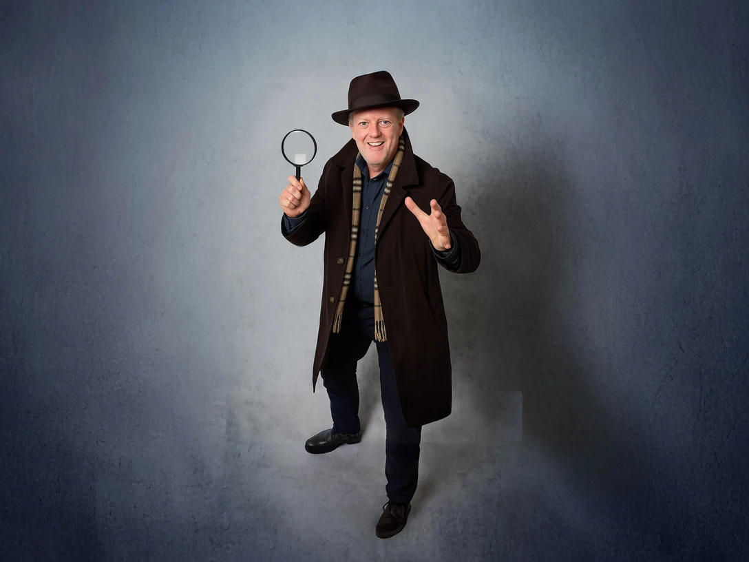 Krimi-Fuehrung-Bad-Zwischenahn.jpg Ein lächelnder Mann in Detektivkleidung mit Lupe, bereit für spannende Erkundungen.