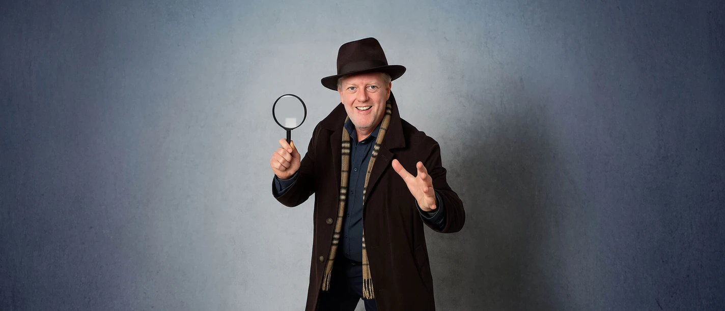 Krimi-Fuehrung-Bad-Zwischenahn.jpg Een lachende man in detectivekleding met een vergrootglas, klaar voor spannende verkenningen.