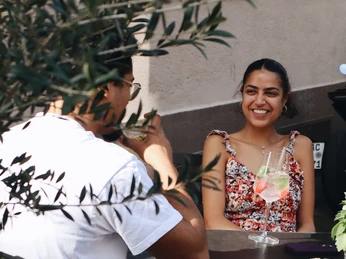 Basic 13 (c) Basic Gastrobar.jpg Zwei Menschen sitzen an einem Tisch im Freien und genießen Getränke. Ein Olivenbaum steht im Vordergrund.