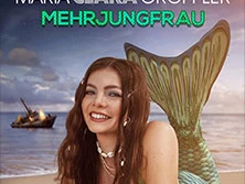 Maria Clara Groppler als Meerjungfrau mit grünem Fischschwanz sitzend vor einem sonnigen Strand.