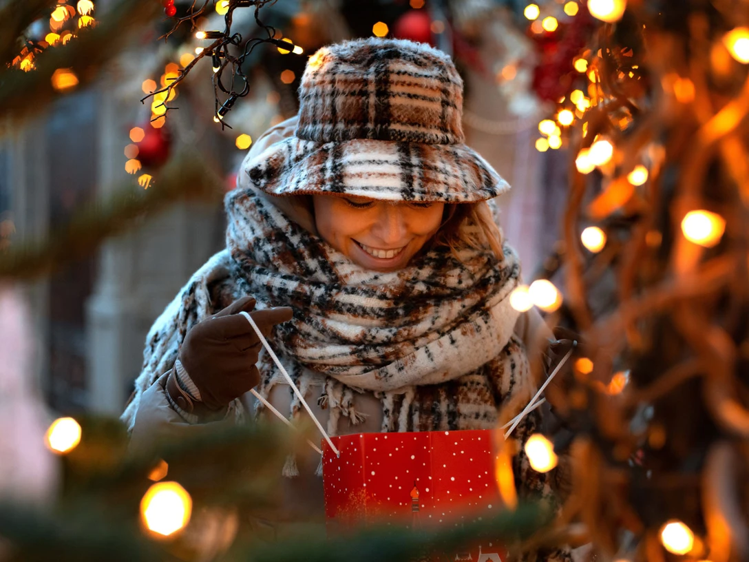 Geschenketausch-Börse Eine junge Frau, warm eingepackt mit Schal und Hut, öffnet gespannt eine rote Geschenktüte.A young woman, wrapped up warmly in a scarf and hat, eagerly opens a red gift bag.En ung kvinde, varmt pakket ind i halstørklæde og hue, åbner ivrigt en rød gavepose.Een jonge vrouw, warm ingepakt in een sjaal en muts, opent gretig een rode geschenktas.