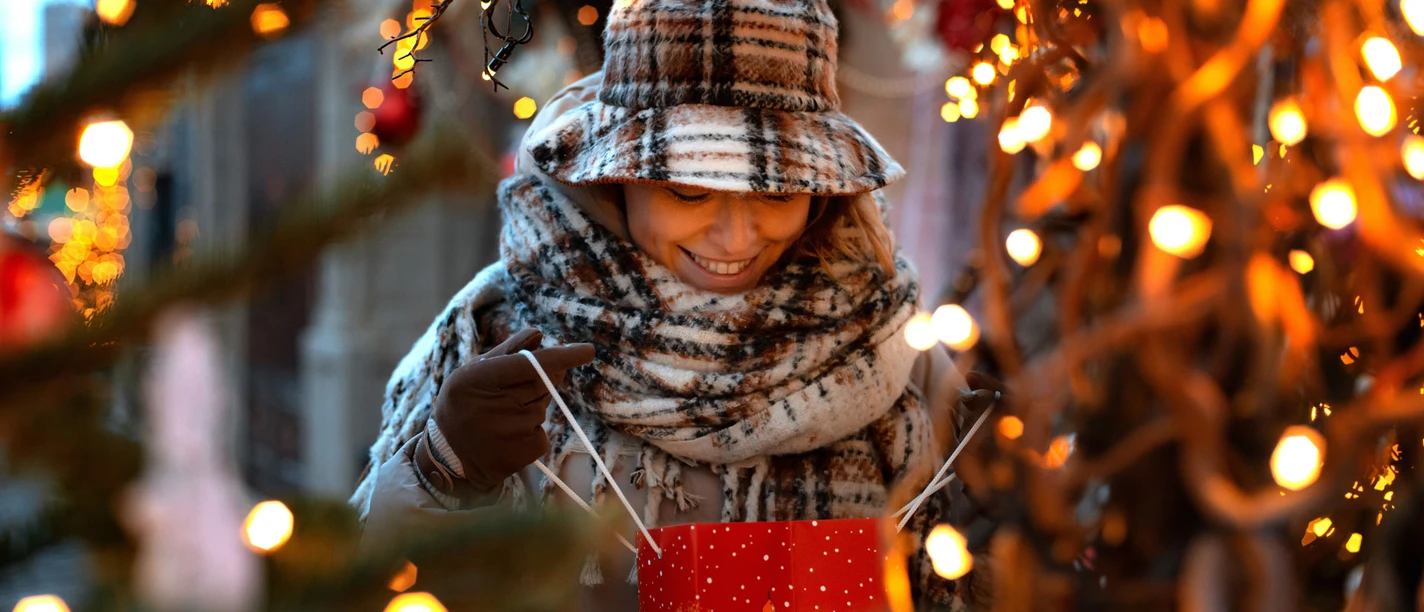 Geschenketausch-Börse Eine junge Frau, warm eingepackt mit Schal und Hut, öffnet gespannt eine rote Geschenktüte.