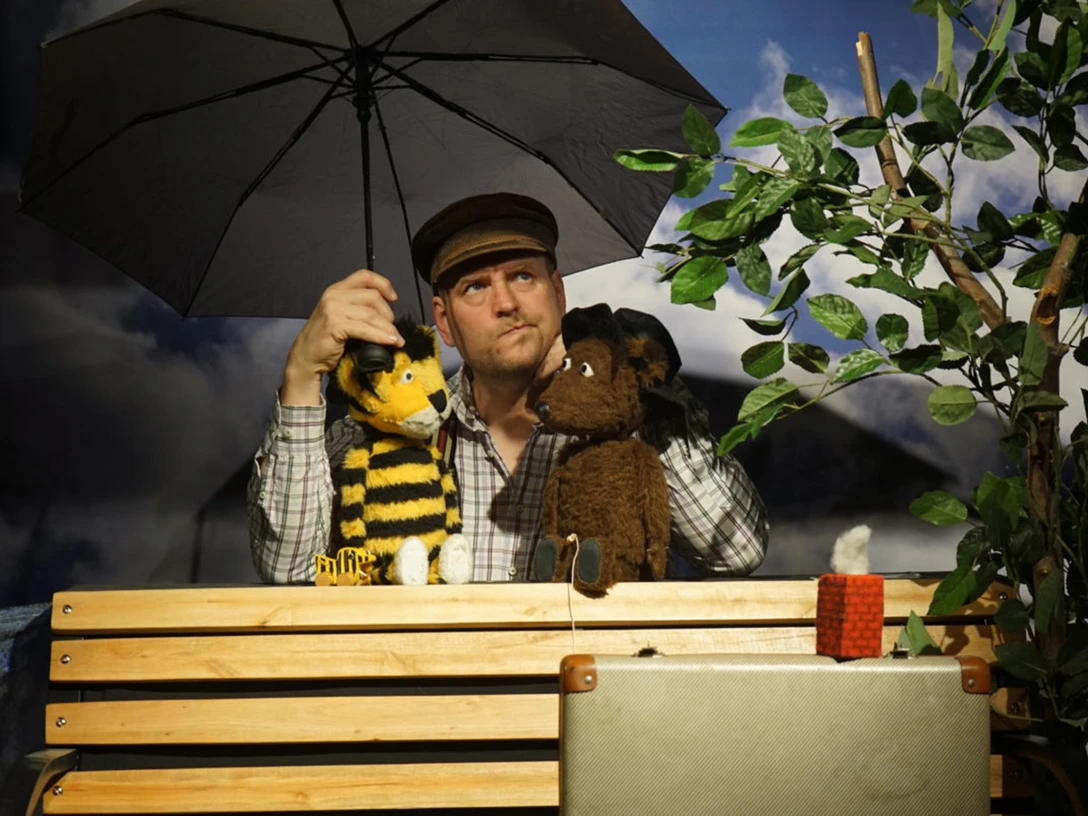 figurentheater-grinsekatze-markus-hesse.jpg Mann mit Schirm spielt Puppentheater mit Biene und Maus auf Bank neben kleinem Baum.