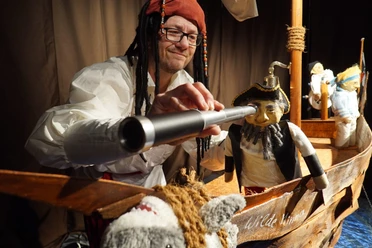 Mann mit Piratenhut blickt durch Fernrohr auf Puppen in einem kleinen hölzernen Piratenschiff.