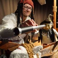 Mann mit Piratenhut blickt durch Fernrohr auf Puppen in einem kleinen hölzernen Piratenschiff.