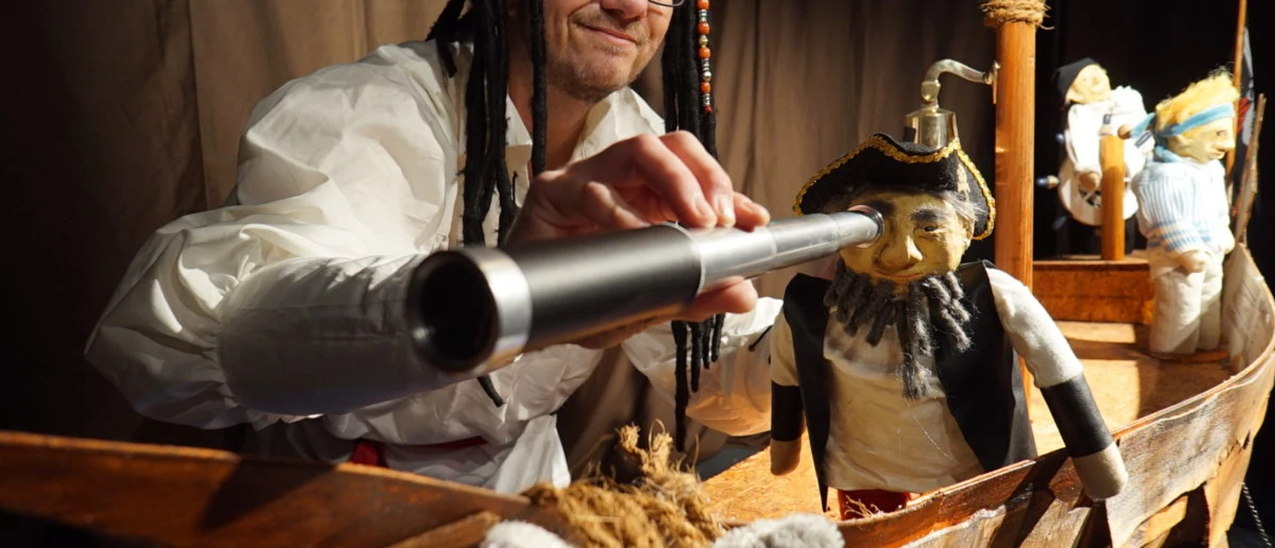Mann mit Piratenhut blickt durch Fernrohr auf Puppen in einem kleinen hölzernen Piratenschiff.