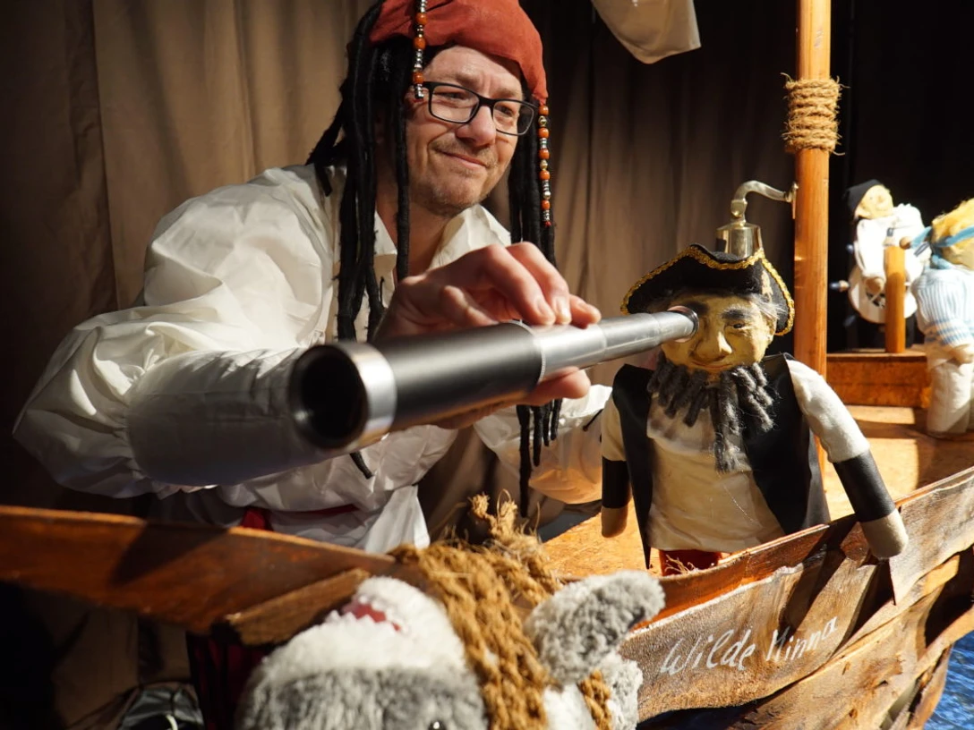 Piratenbild Mann mit Piratenhut blickt durch Fernrohr auf Puppen in einem kleinen hölzernen Piratenschiff.
