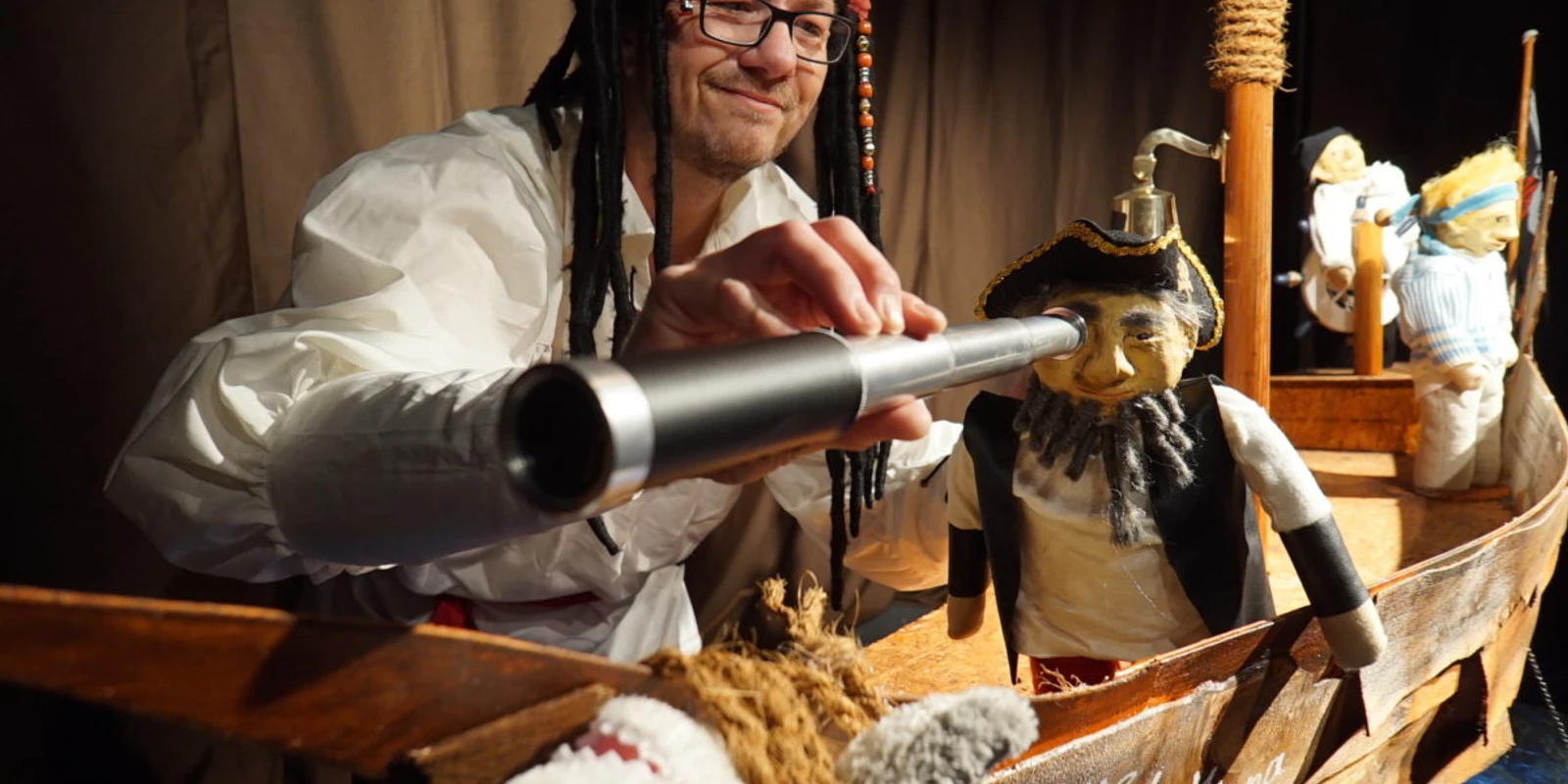Piratenbild Mann mit Piratenhut blickt durch Fernrohr auf Puppen in einem kleinen hölzernen Piratenschiff.