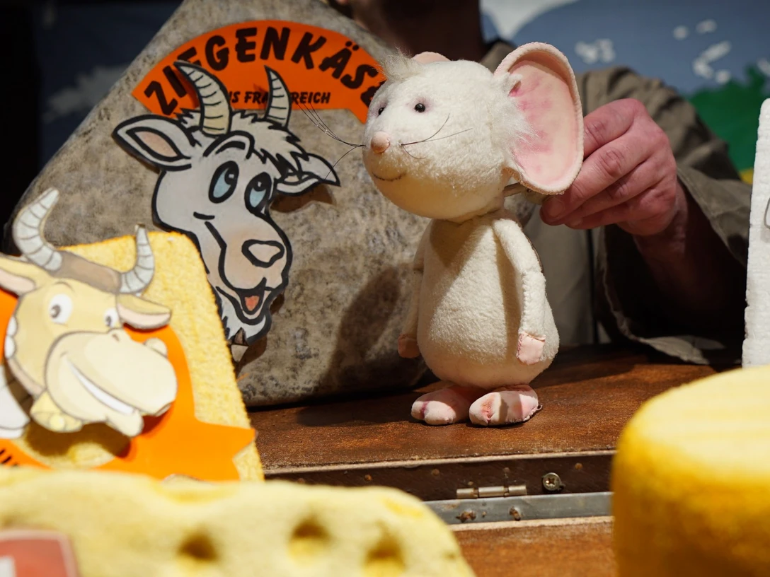 figurentheater-grinsekatze-liebe-gruesse-deine-tilda-1.jpg Eine weiße Stoffmaus auf einer Bühne zwischen großen Käseattrappen beim Figurentheater.