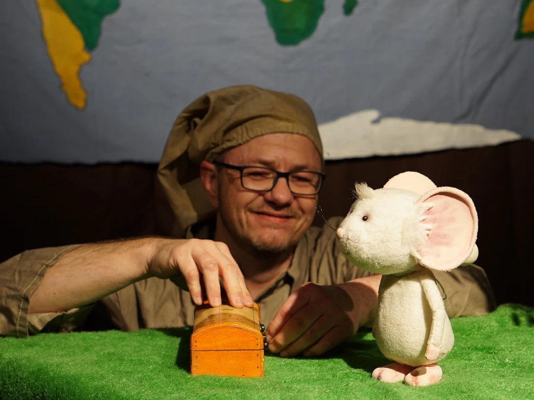 figurentheater-grinsekatze-liebe-gruesse-deine-tilda-2.jpg Puppenspieler mit Schlafmütze spielt auf grüner Bühne mit weißer Mausfigur und kleiner Holzkiste.