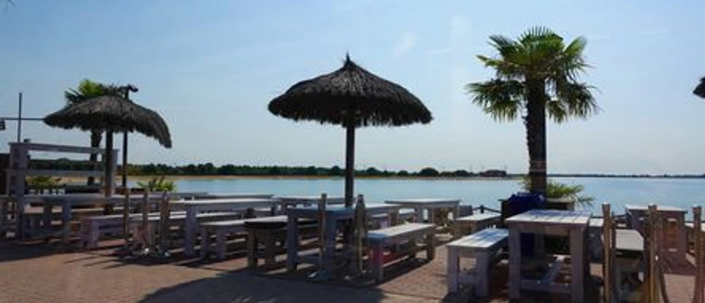 Strandcafé mit Holztischen, Palmen und Sonnenschirmen am Ufer eines klaren Sees unter blauem Himmel.