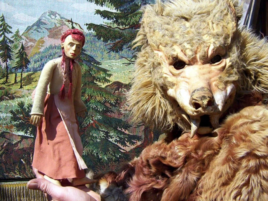 figurentheater-ernst-heiter-rotkaeppchen.jpg Holzfigur Rotkäppchen und wolfsähnliche Maske vor gemaltem Waldhintergrund einer Theaterbühne