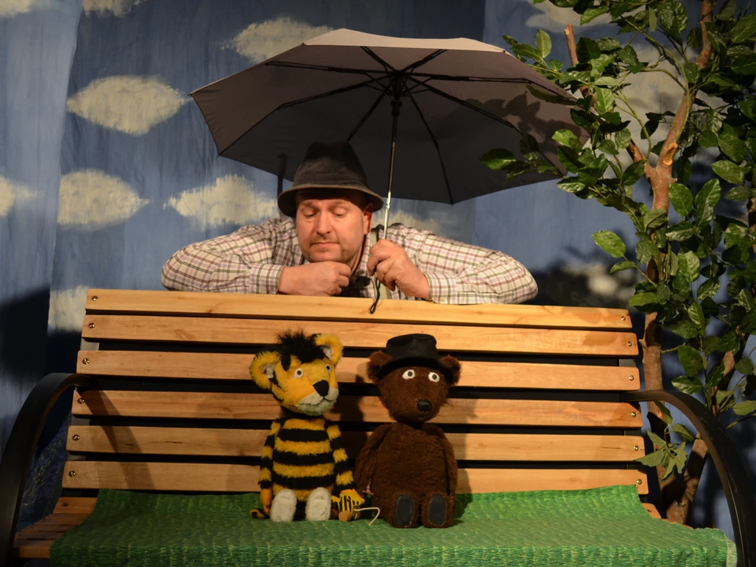 figurentheater-grinsekatze-oh-wie-schoen-ist-panama-1.jpg Mann mit Regenschirm hinter zwei Puppen, Tiger und Bär, auf Bank vor blauem Himmelhintergrund