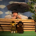 figurentheater-grinsekatze-oh-wie-schoen-ist-panama-1.jpg Mann mit Regenschirm hinter zwei Puppen, Tiger und Bär, auf Bank vor blauem Himmelhintergrund