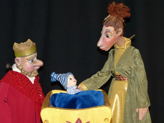 annes-buehne-der-prinz-mit-den-eselsohren-1.jpg Figurenpuppen eines Königs, einer Königin und eines Babys in Wiege vor dunklem Hintergrund