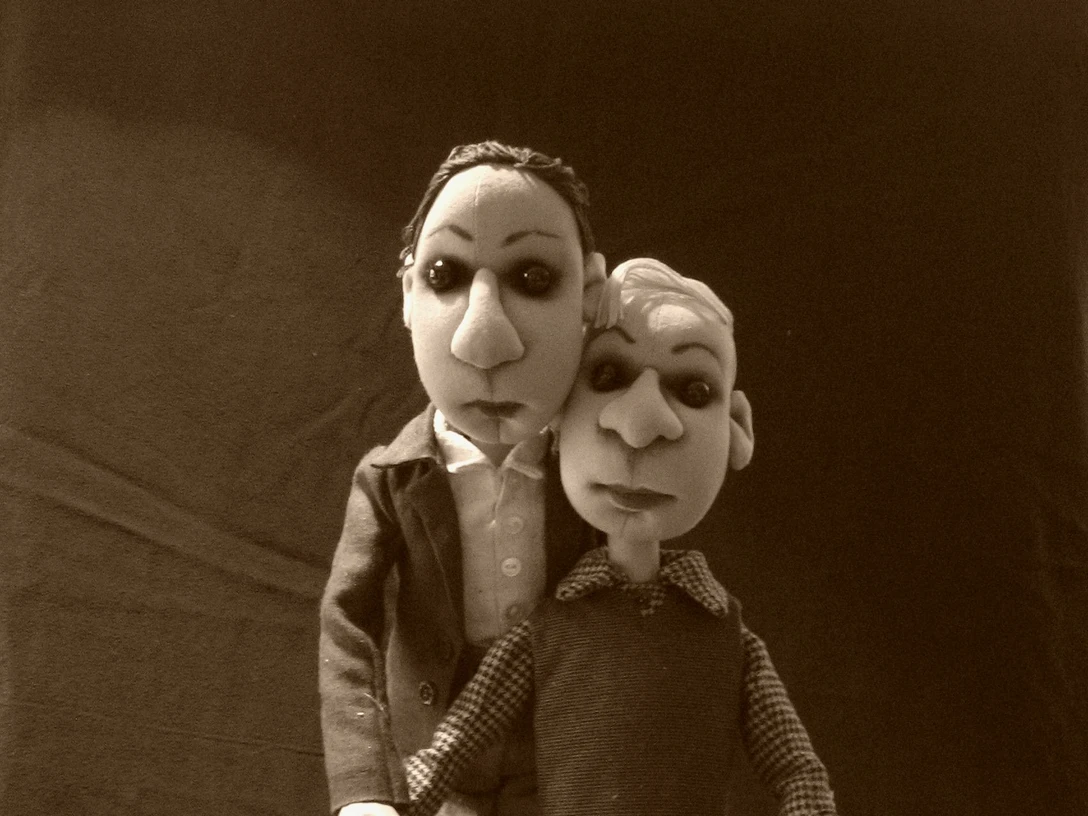 seifenblasen-theater-hannes-und-paul.jpg Zwei Puppenfiguren sitzen eng auf einer Kiste und blicken ernst in die Kamera.