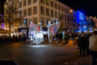 2024_11_02_Moonlight_Shopping_©HolgerBulk-169.jpg Zwei kostümierte Stelzenkünstler mit leuchtenden Flügeln erhellen eine Einkaufsstraße bei Nacht.