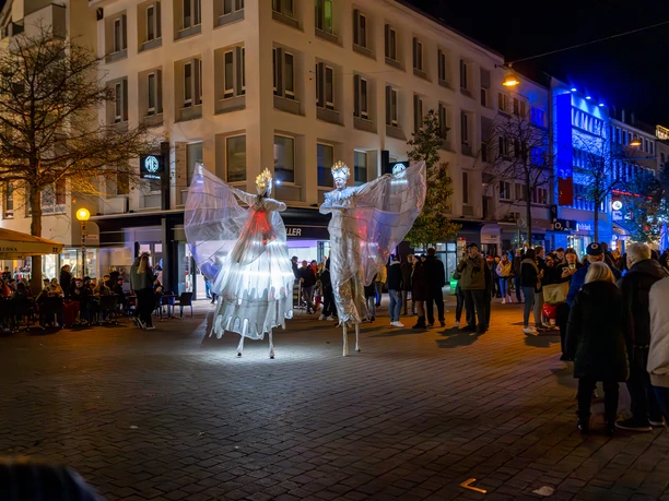 2024_11_02_Moonlight_Shopping_©HolgerBulk-169.jpg Zwei kostümierte Stelzenkünstler mit leuchtenden Flügeln erhellen eine Einkaufsstraße bei Nacht.