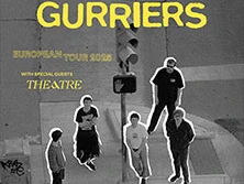 Fünf Personen stehen auf einem Bürgersteig bei einer Fußgängerampel, im Hintergrund Straßenkreuzung.