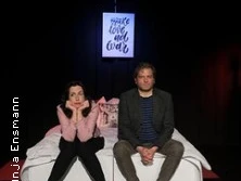 Zwei Personen sitzen auf einem Bett in einem dunklen Raum, ein Poster hängt mit der Aufschrift "Make Love Not War".