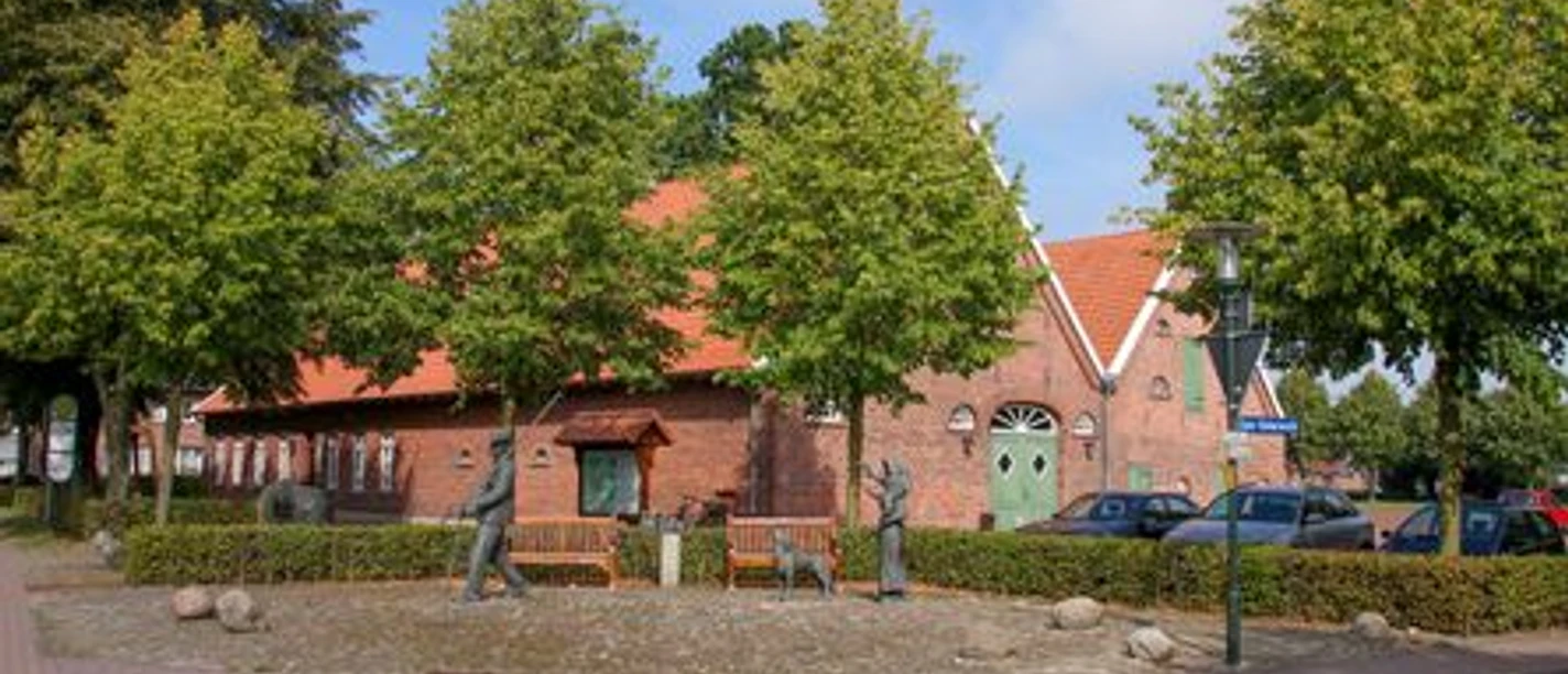 Backsteingebäude mit rotem Dach, umgeben von Bäumen, Sitzbänken und kleinen Bronzefiguren.