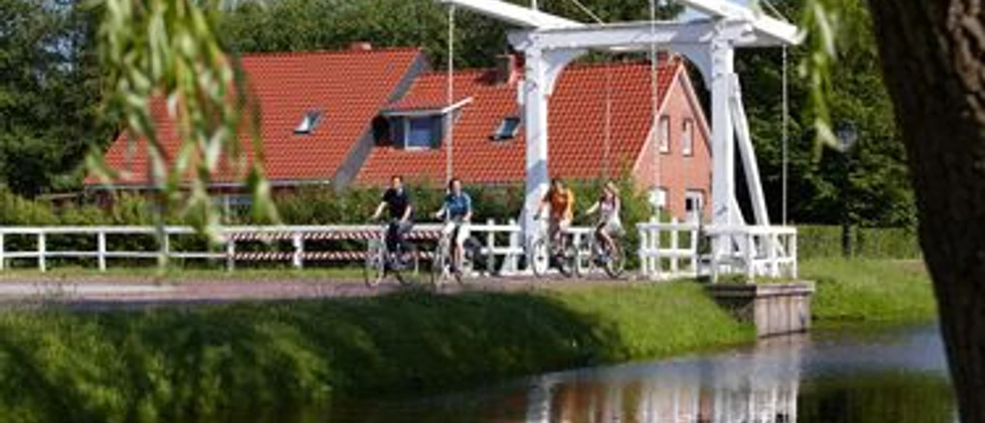 Radfahrer auf einer weißen Klappbrücke über einem Kanal vor roten Ziegeldächern in Papenburg