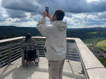 Rollstuhl Ein Mann fotografiert die Landschaft, ein Rollstuhlfahrer blickt von der Aussicht in die Landschaft.