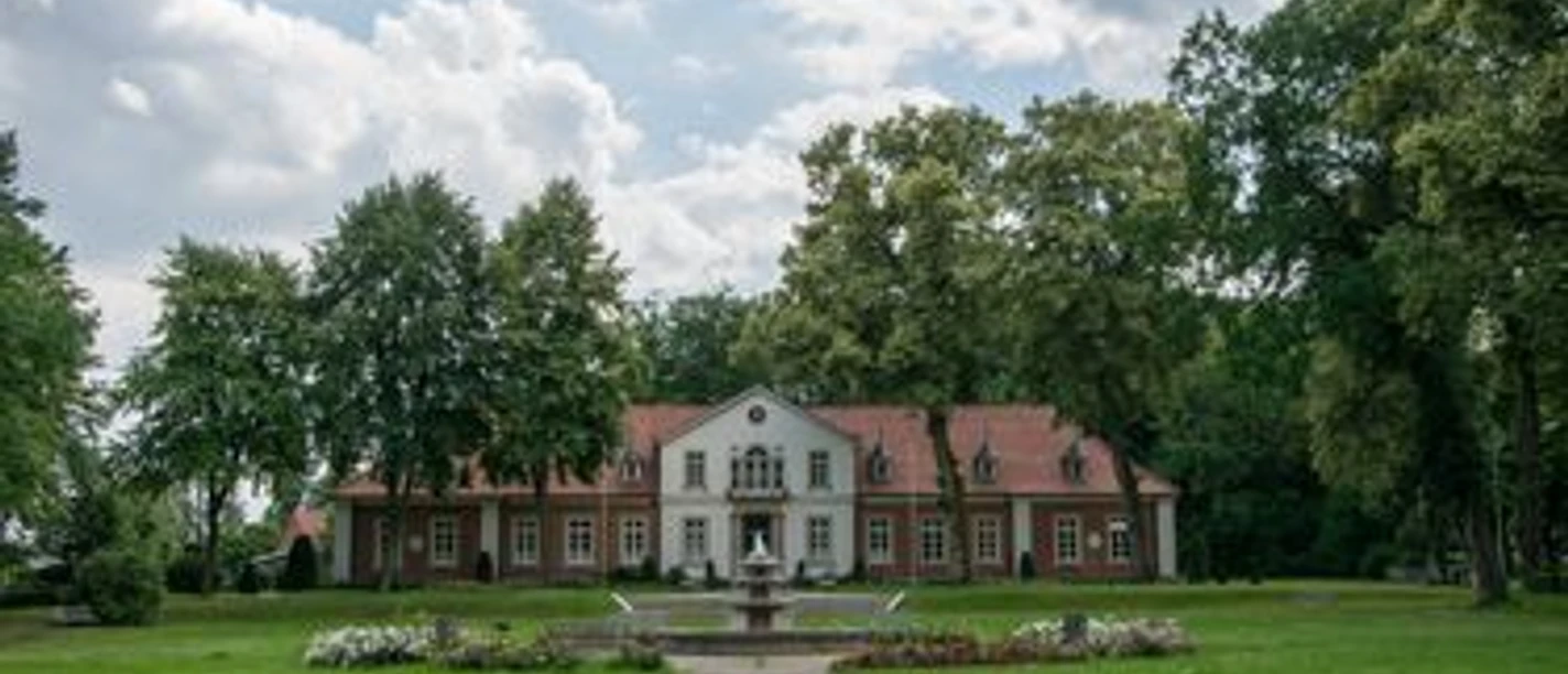 Historisches Gebäude mit rotem Dach und Brunnen im Park von Sögel, umgeben von alten Bäumen