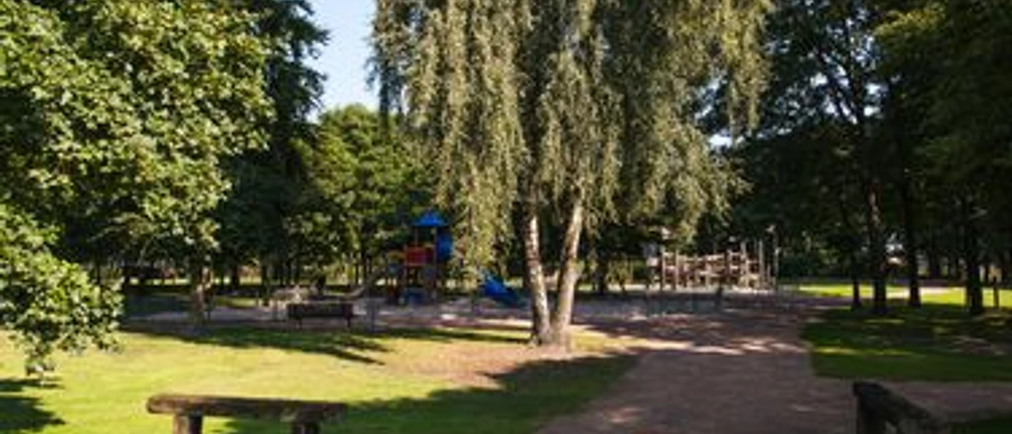 Grüner Park mit schattigen Bäumen, Holzbänken und einem großen Spielplatz im Hintergrund.