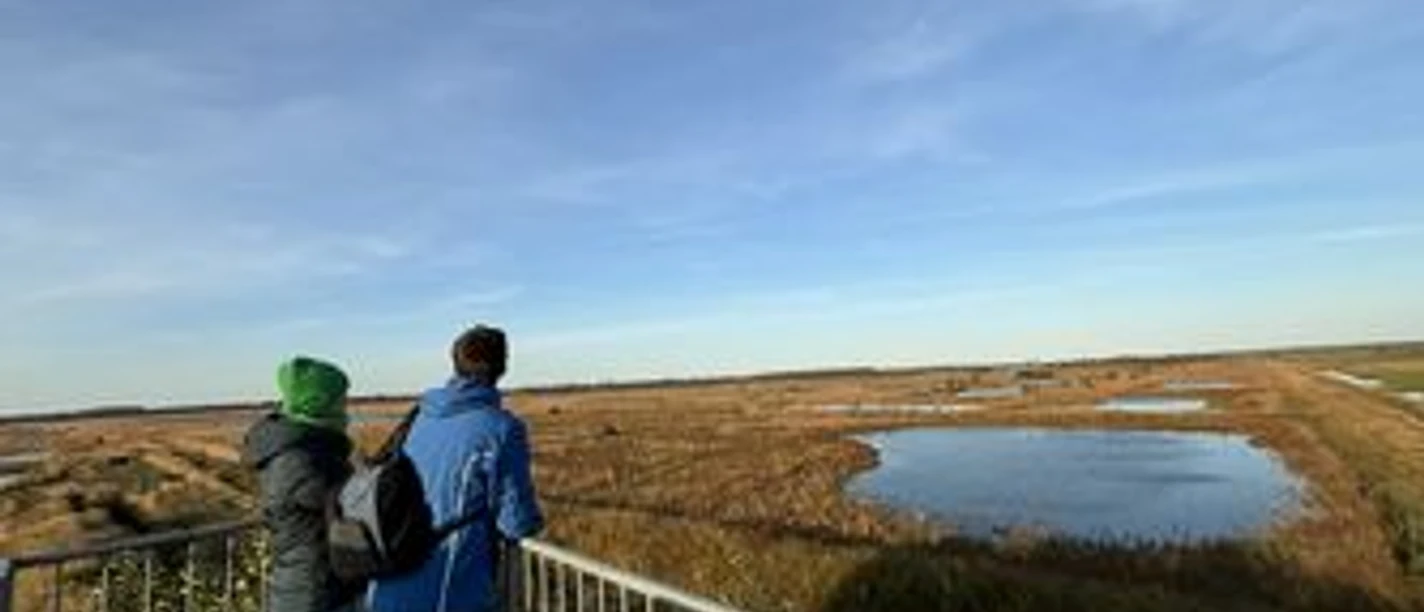 Zwei Menschen blicken von einer Aussichtsplattform über eine weite Moorlandschaft mit kleinem See.